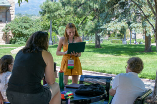 A VCFA MFA in Writing for Children & Young Adults alumnx guides an outdoor discussion with current students at summer residency in Colorado. She holds her laptop and smiles as the students around her listen intently.