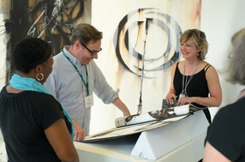 The VCFA Academic Dean smiles as he looks through a student's graphic design portfolio during a residency critique. Two women smile at each other as the Dean examines the artwork.