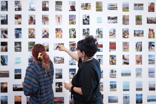 Alt-text: A VCFA faculty member and a student examine a gallery wall filled with evenly-spaced photographs, and the faculty member points something out to the student.