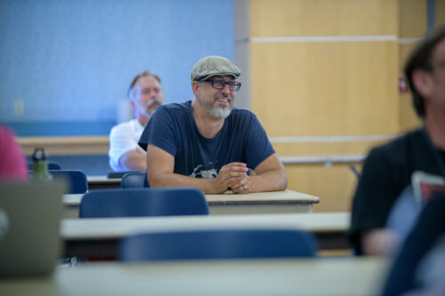 An instructor sits at a desk in a classroom full of students, smiling as he listens to the presenter, who is off-screen.