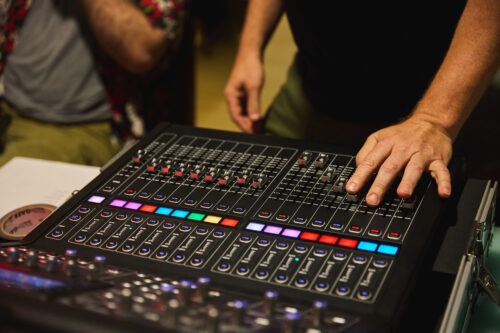 A close-up image of a sound board with multicolored buttons. The musician is out of view, except for their hands.