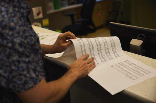 A VCFA student stands at a table, reading a longer piece of sheet music. The student is primarily out of view, and only their arms are visible.