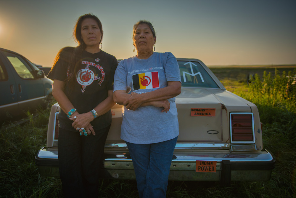 A Native American mother and daughter stand in front of a car and face the camera with a powerful gaze